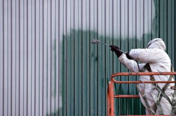 Worker in protective suit spray-painting a corrugated metal wall green from an orange lift platform